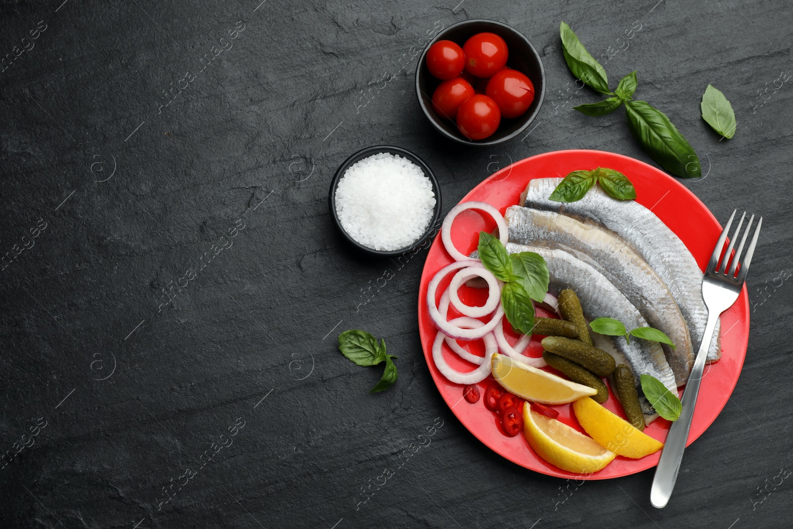 Photo of Salted herring fillets served with onion rings, pickles, basil, chili pepper and lemon on black table, flat lay. Space for text