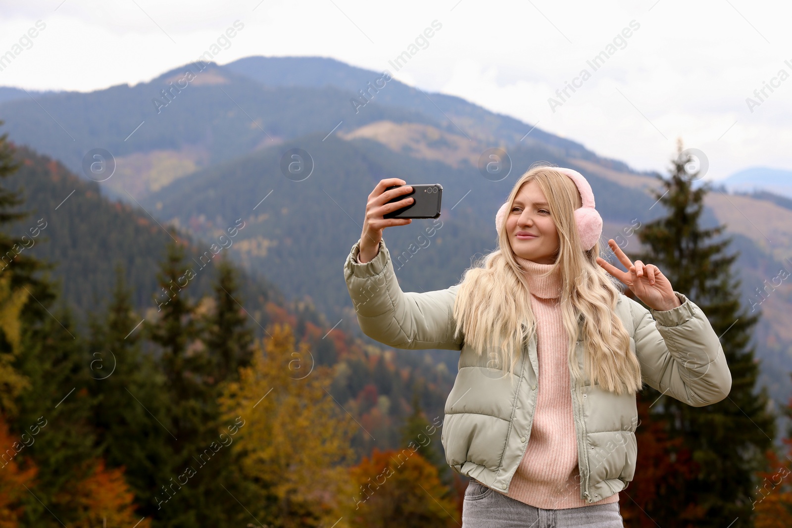 Photo of Young woman in warm earmuffs taking selfie on autumn day