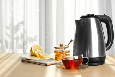 Photo of Modern electric kettle and cup of tea on wooden table indoors. Space for text