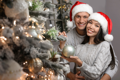 Photo of Happy young couple in Santa hats decorating Christmas tree together at home