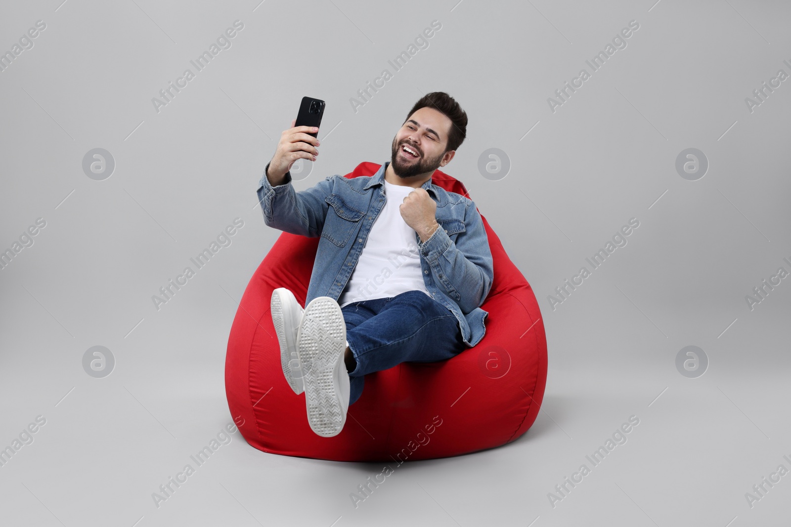Photo of Happy young man using smartphone on bean bag chair against grey background