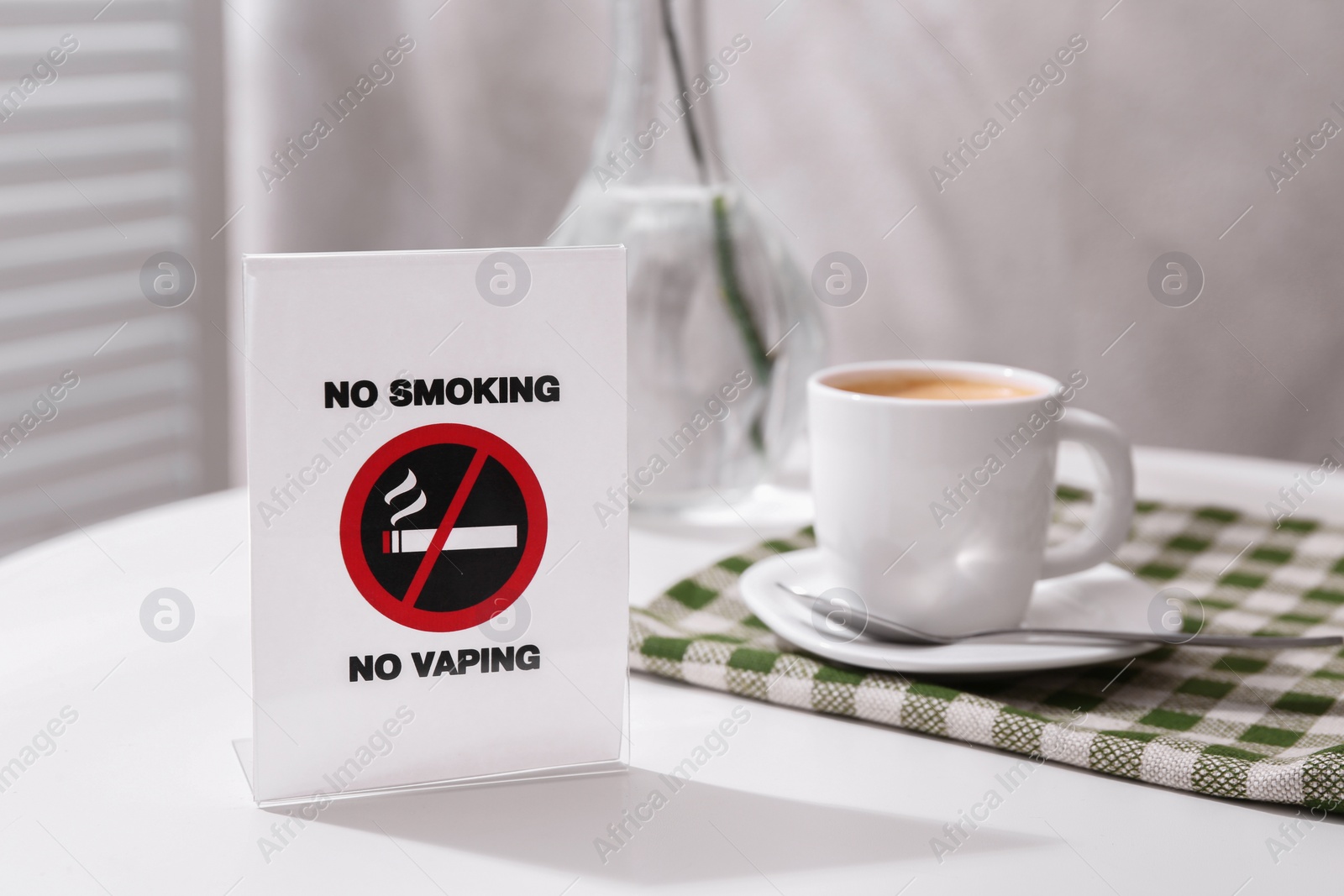 Photo of No Smoking No Vaping sign and cup of coffee on white table indoors