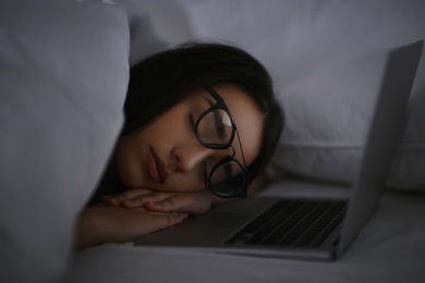 Photo of Young woman with nomophobia sleeping near laptop in bed at night