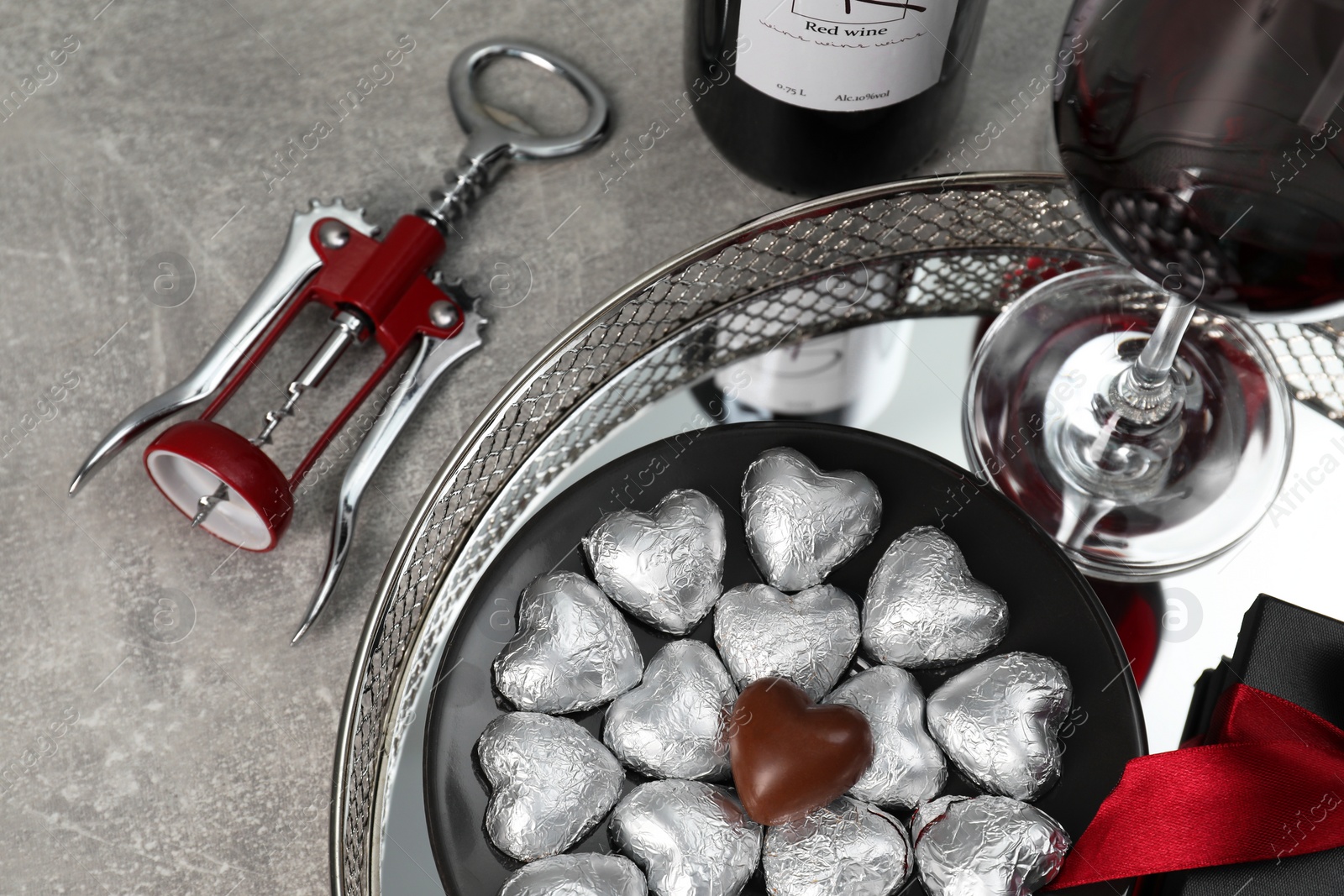 Photo of Bottle, glass of red wine, heart shaped chocolate candies, corkscrew and gift box on light grey textured table, above view