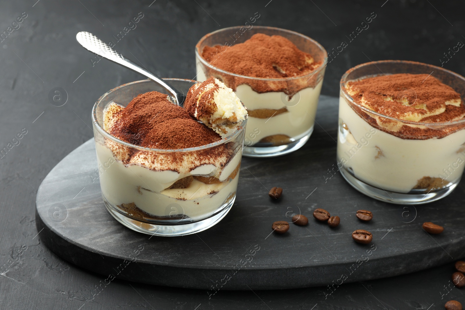 Photo of Delicious tiramisu in glasses, spoon and coffee beans on black table