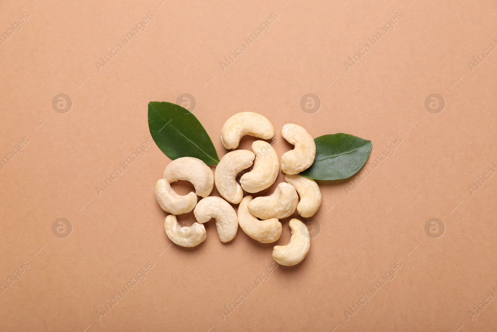 Photo of Pile of tasty cashew nuts and green leaves on pale brown background, flat lay