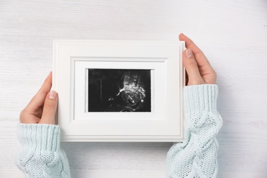 Photo of Woman holding ultrasound photo of baby over wooden table, top view