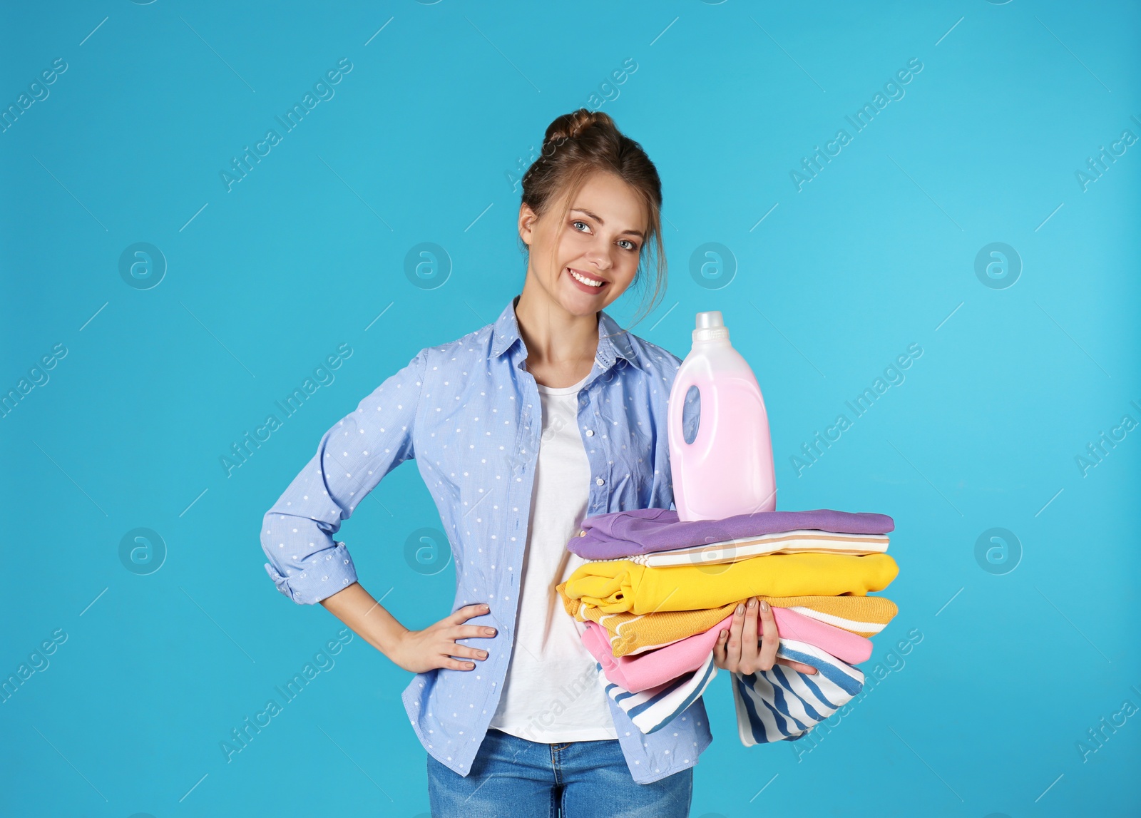 Photo of Happy young woman holding clean clothes and laundry detergent on color background