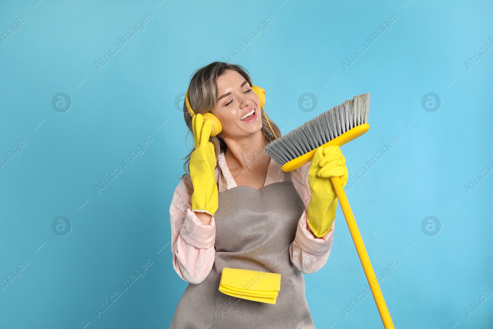 Photo of Beautiful young woman with headphones and floor brush singing on light blue background