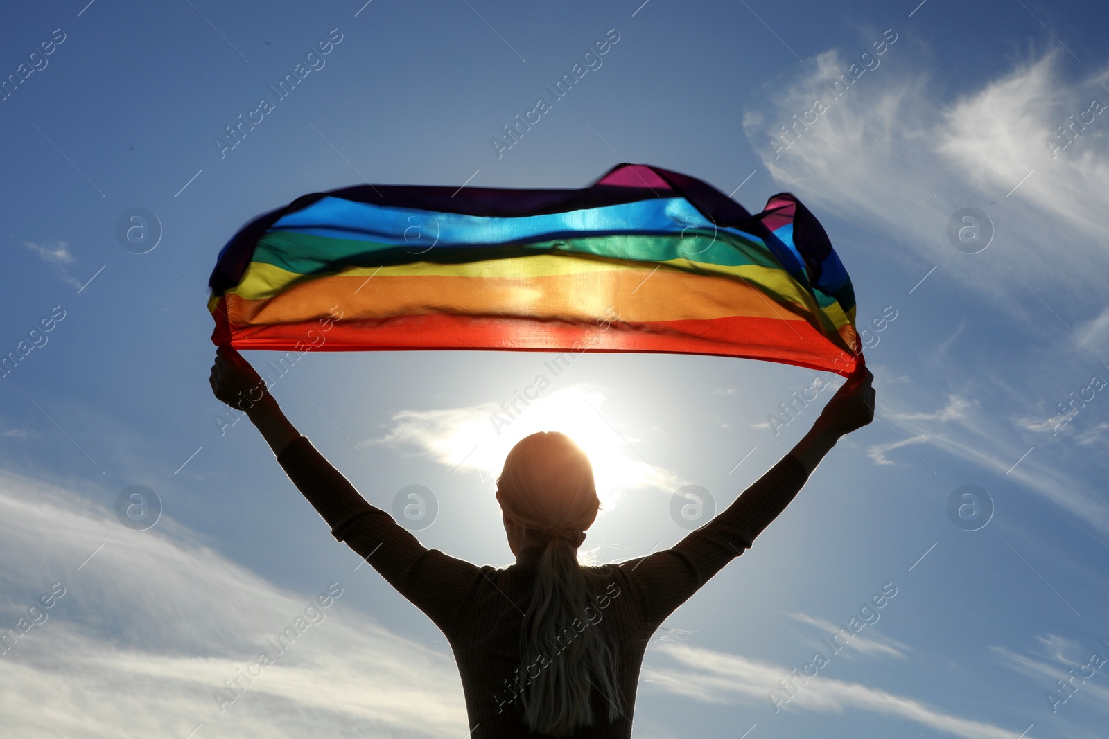 Photo of Woman holding bright LGBT flag against blue sky on sunny day, back view