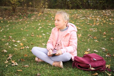 Photo of Cute little girl with copybook and backpack on green grass outdoors