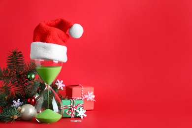 Photo of Hourglass with Santa hat and decor on red background, space for text. Christmas countdown