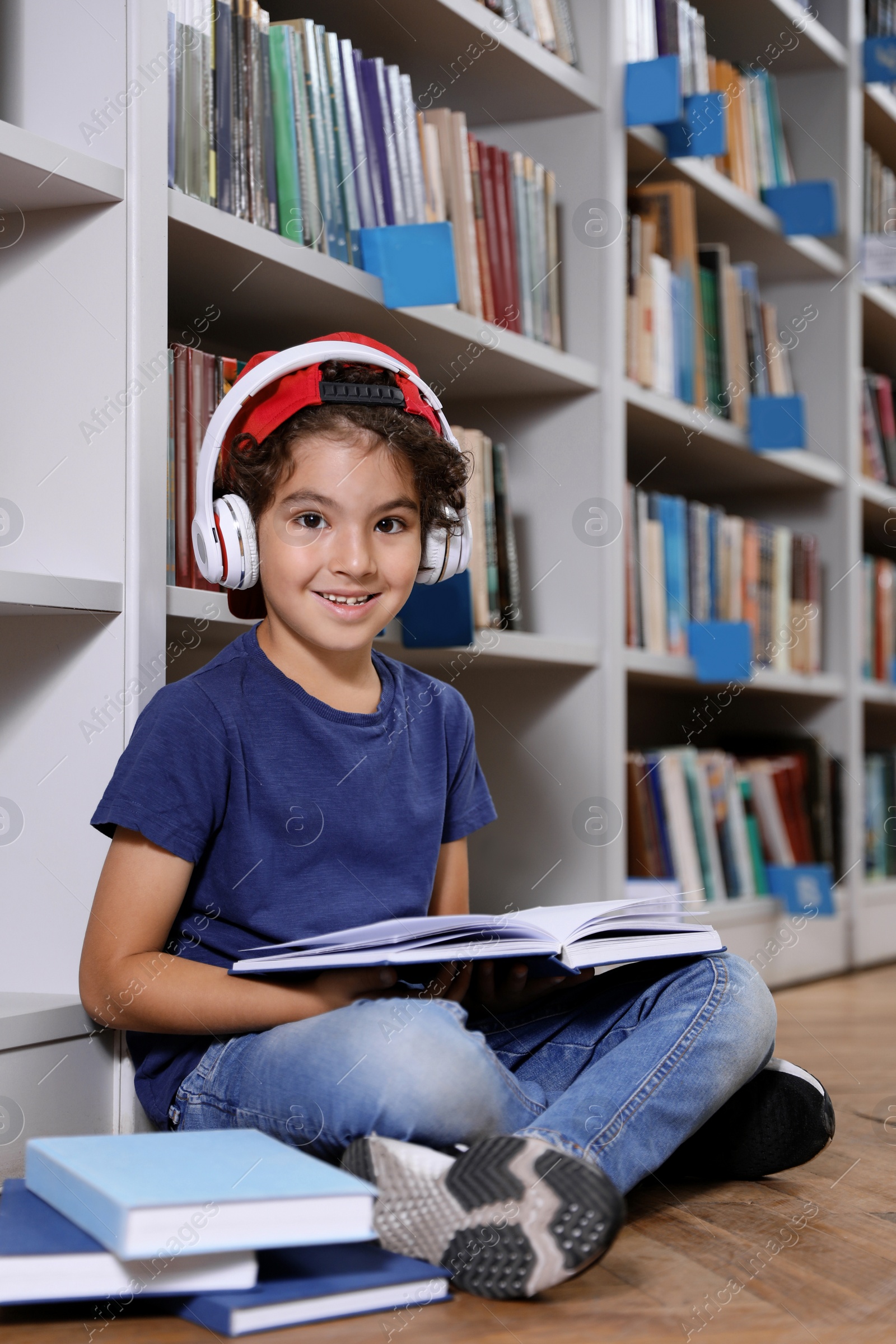 Photo of Cute little boy with headphones reading book on floor in library