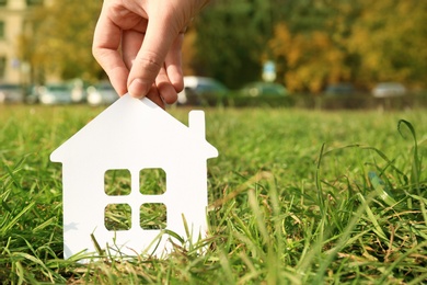 Photo of Woman holding paper silhouette of house in grass outdoors, space for text. Insurance concept