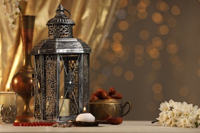 Photo of Arabic lantern, misbaha, candles, dates and flowers on table against blurred lights