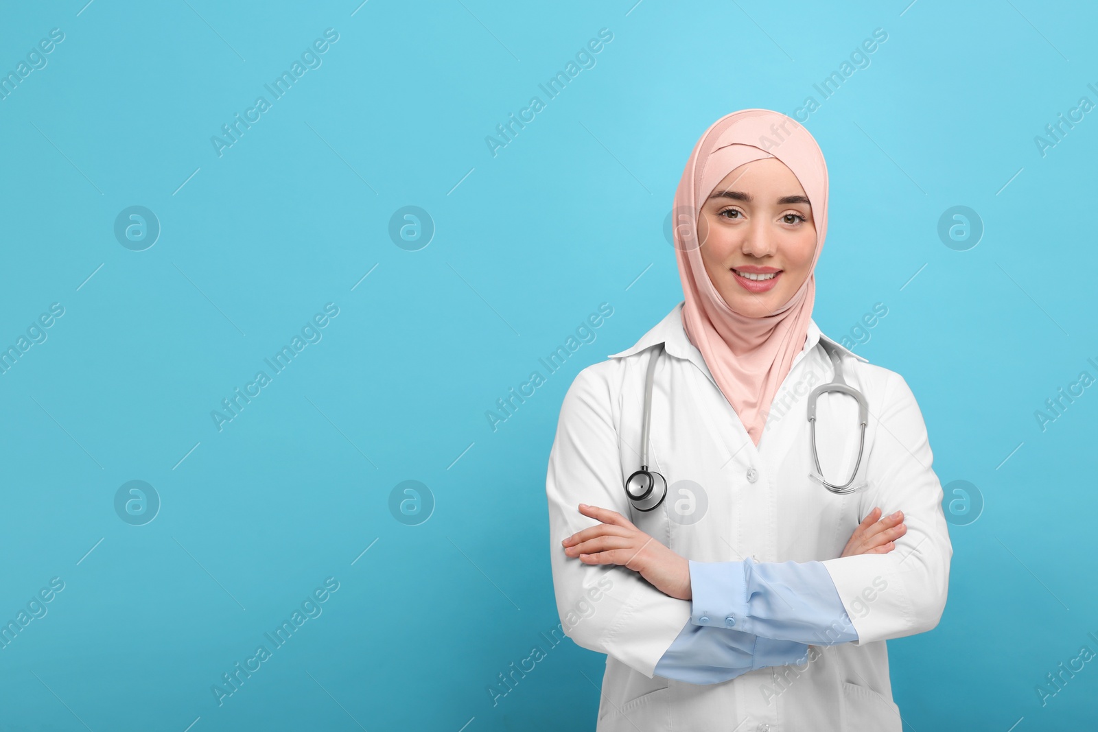 Photo of Muslim woman wearing hijab and medical uniform with stethoscope on light blue background, space for text