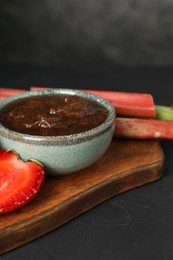 Tasty rhubarb jam in bowl, stems and strawberry on dark table