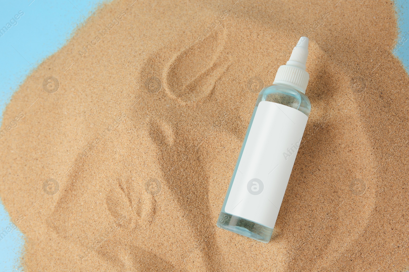 Photo of Bottle with serum on sand against light blue background, top view and space for text. Cosmetic product