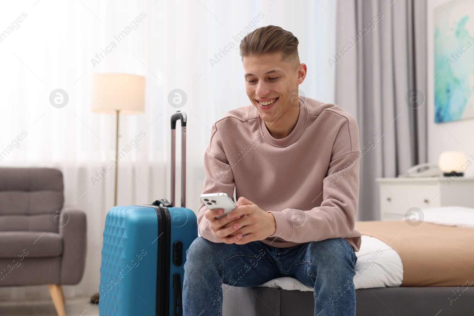 Photo of Smiling guest with smartphone relaxing on bed in stylish hotel room