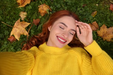 Smiling woman lying on grass among autumn leaves and taking selfie, top view