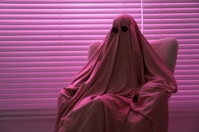 Photo of Glamorous ghost. Woman in sheet and sunglasses on armchair in pink light, space for text