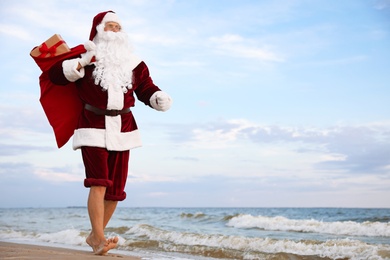 Photo of Santa Claus with bag of presents on beach, space for text. Christmas vacation