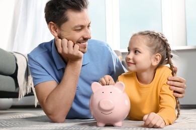 Photo of Little girl with her father putting coin into piggy bank at home