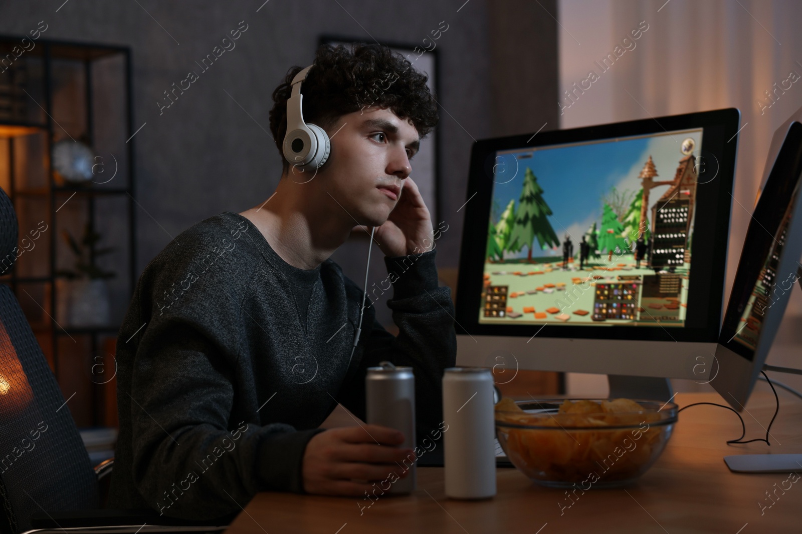 Photo of Young man with energy drink and headphones playing video game at wooden desk indoors