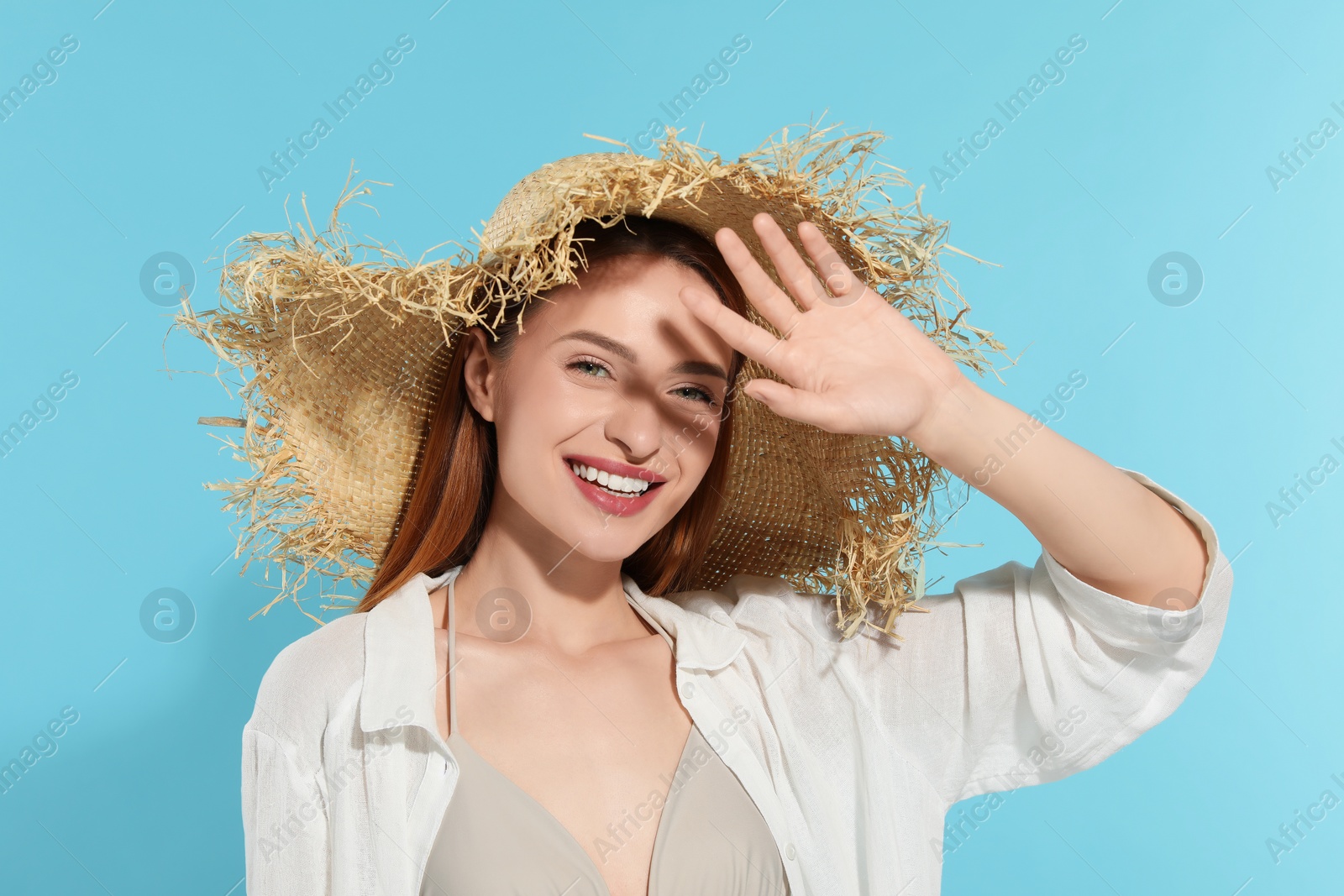 Photo of Beautiful young woman in straw hat shading herself with hand from sunlight on light blue background