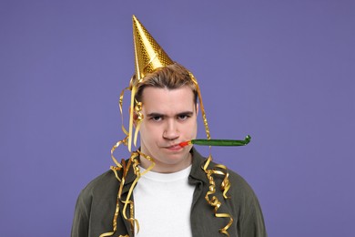 Sad young man with party hat and blower on purple background