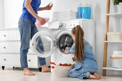 Photo of Mother pouring fabric softener and daughter putting dirty clothes into washing machine in bathroom, closeup