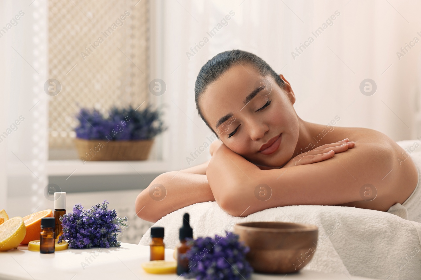 Photo of Beautiful young woman relaxing on massage couch and bottles of essential oil with ingredients on table in spa salon