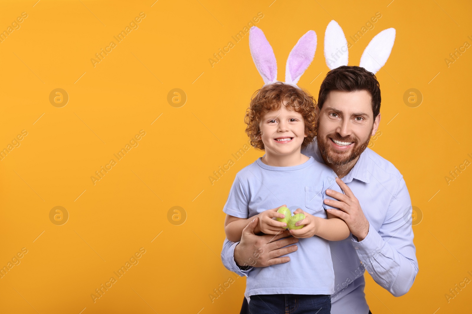 Photo of Happy father and son wearing cute bunny ears headbands on orange background. Boy holding Easter eggs, space for text