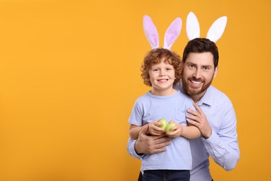 Photo of Happy father and son wearing cute bunny ears headbands on orange background. Boy holding Easter eggs, space for text