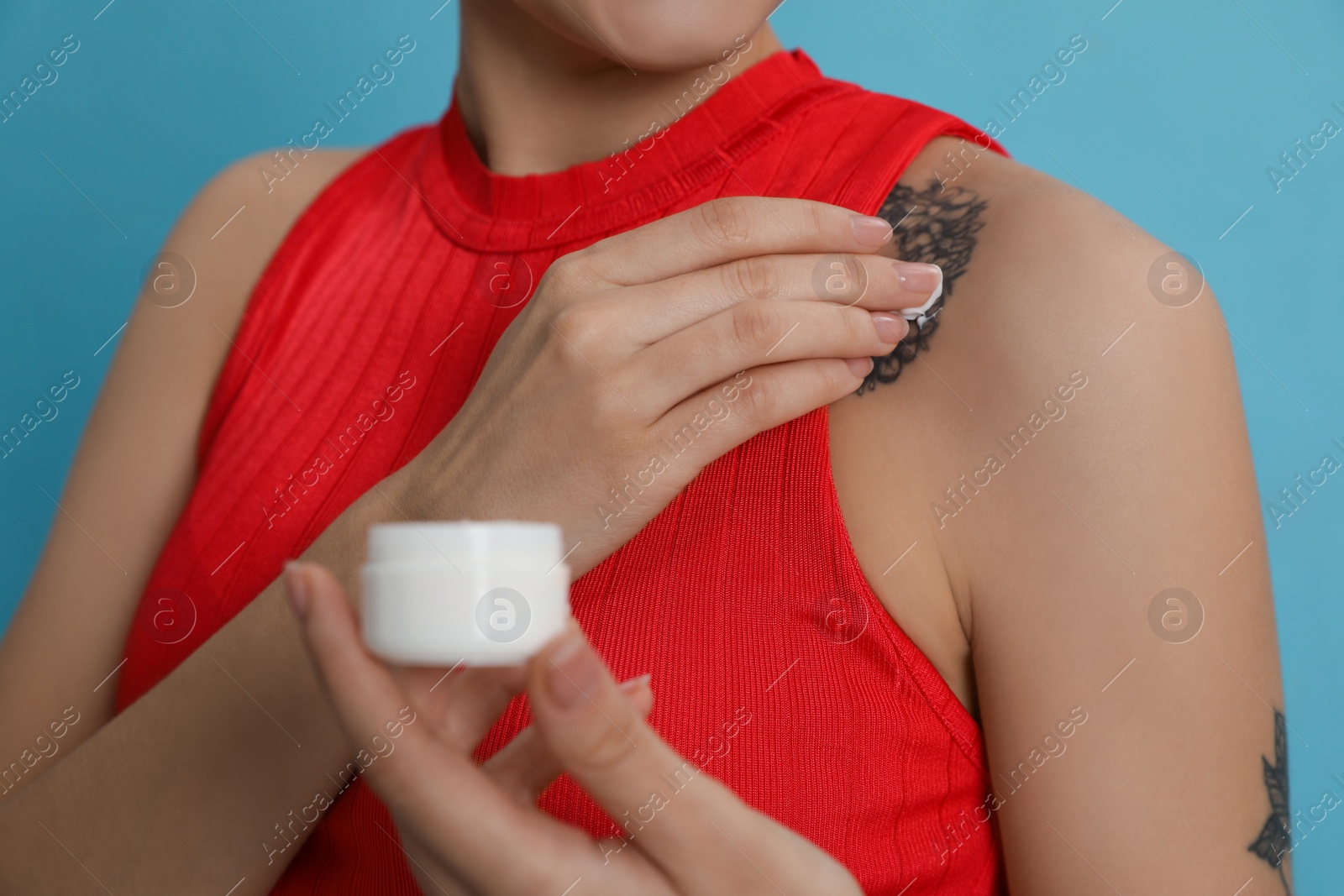 Photo of Woman applying cream onto tattoo on her skin against light blue background, closeup