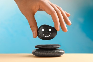 Photo of Woman putting stone with drawn happy face on stack against light blue background, closeup. Zen concept