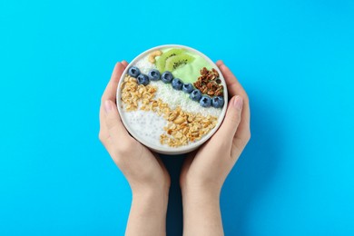 Photo of Woman holding tasty smoothie bowl with fresh kiwi fruit, blueberries and oatmeal at light blue table, top view