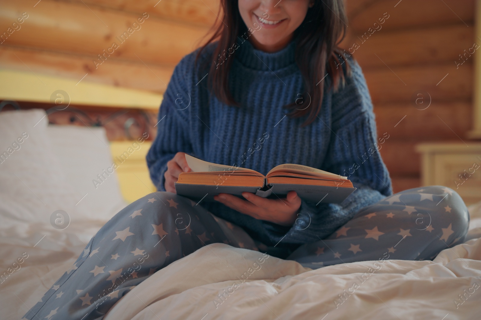 Photo of Young woman in warm sweater reading book on bed at home, closeup