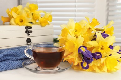 Photo of Cup of aromatic tea, beautiful yellow daffodil and iris flowers on windowsill