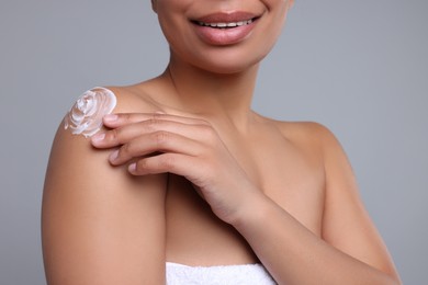 Young woman applying body cream onto shoulder on grey background, closeup
