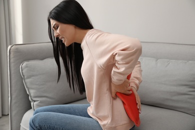Photo of Woman using hot water bottle to relieve low back pain on sofa at home