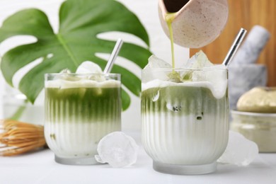 Pouring tasty matcha tea into glasses with milk and ice cubes at white table, closeup