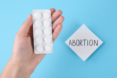 Photo of Woman holding pills near paper note with word Abortion on light blue background, top view