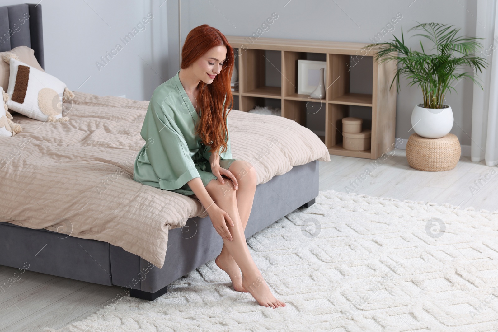 Photo of Beautiful young woman touching her smooth legs in bedroom, space for text