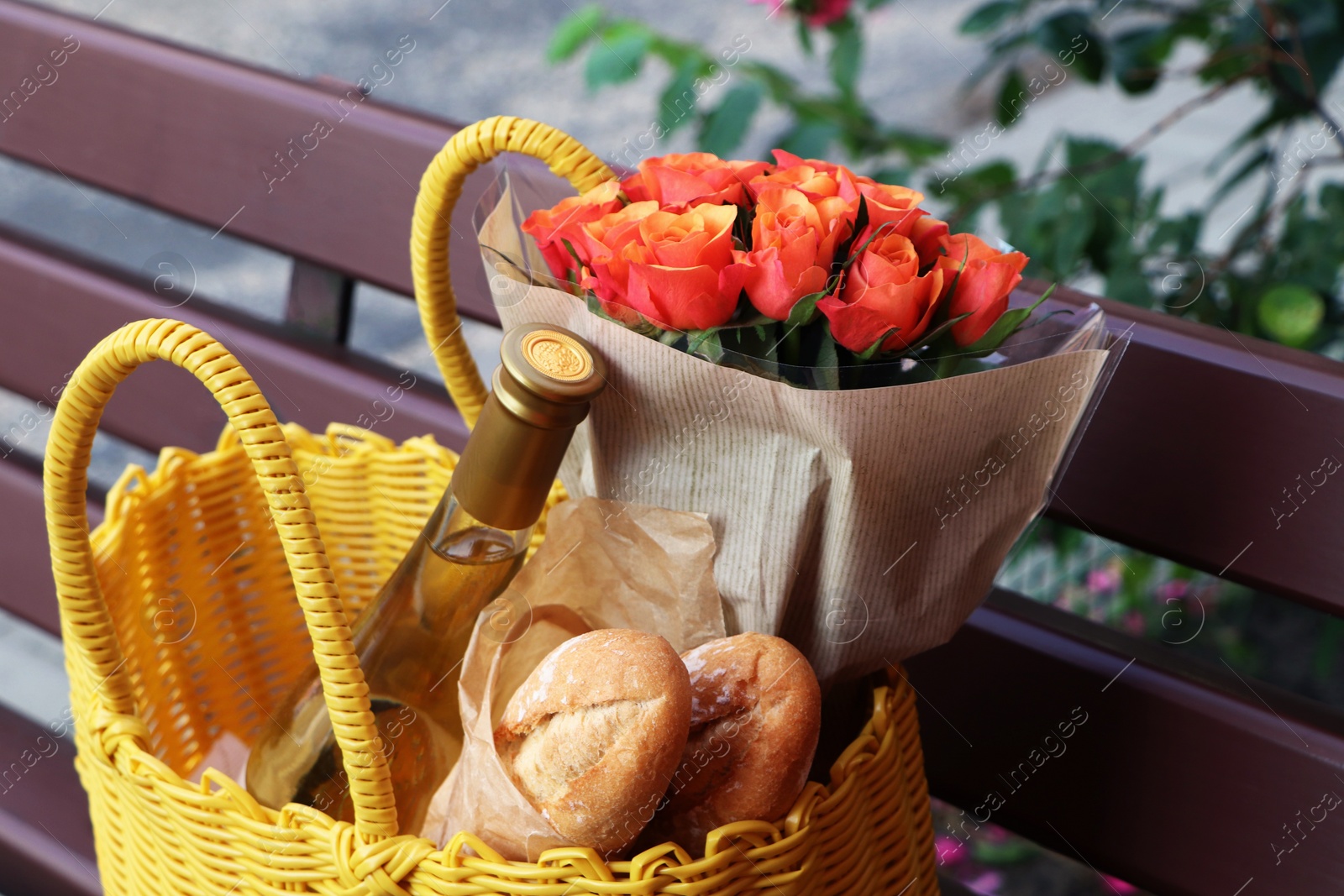 Photo of Yellow wicker bag with beautiful roses, bottle of wine and baguettes on bench outdoors, closeup