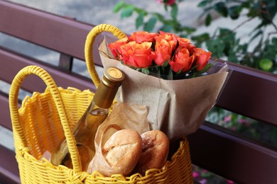 Photo of Yellow wicker bag with beautiful roses, bottle of wine and baguettes on bench outdoors, closeup