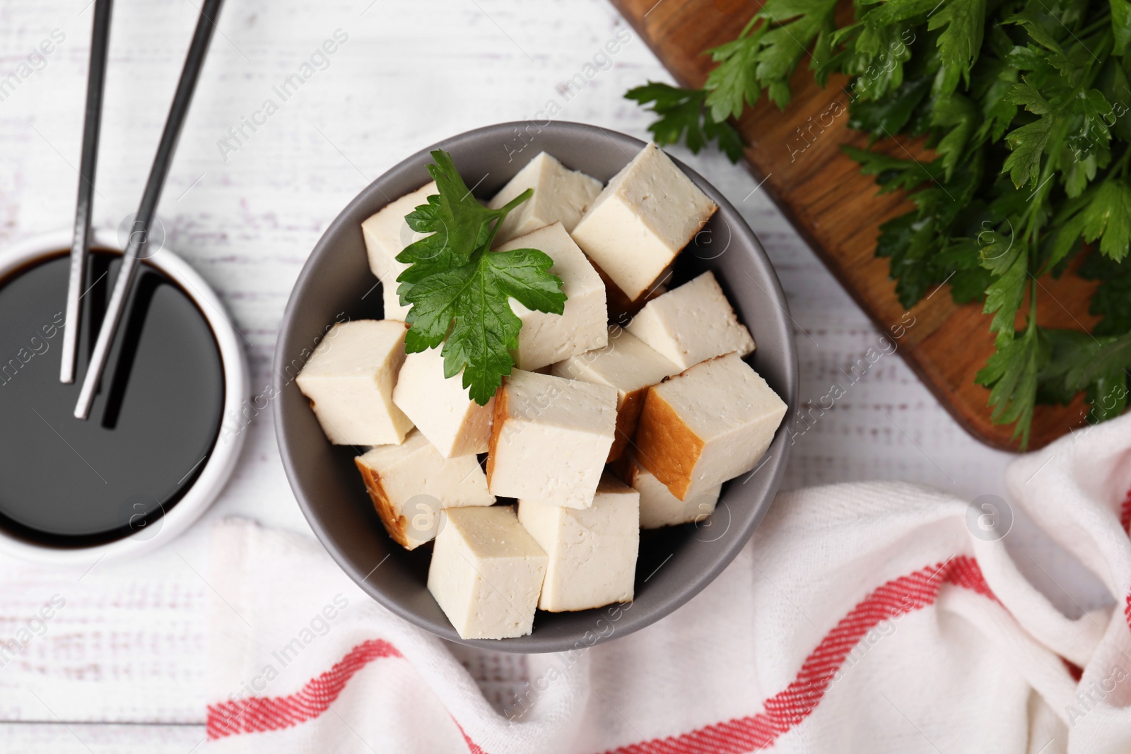 Photo of Bowl of smoked tofu cubes, soy sauce and parsley on white wooden table, flat lay