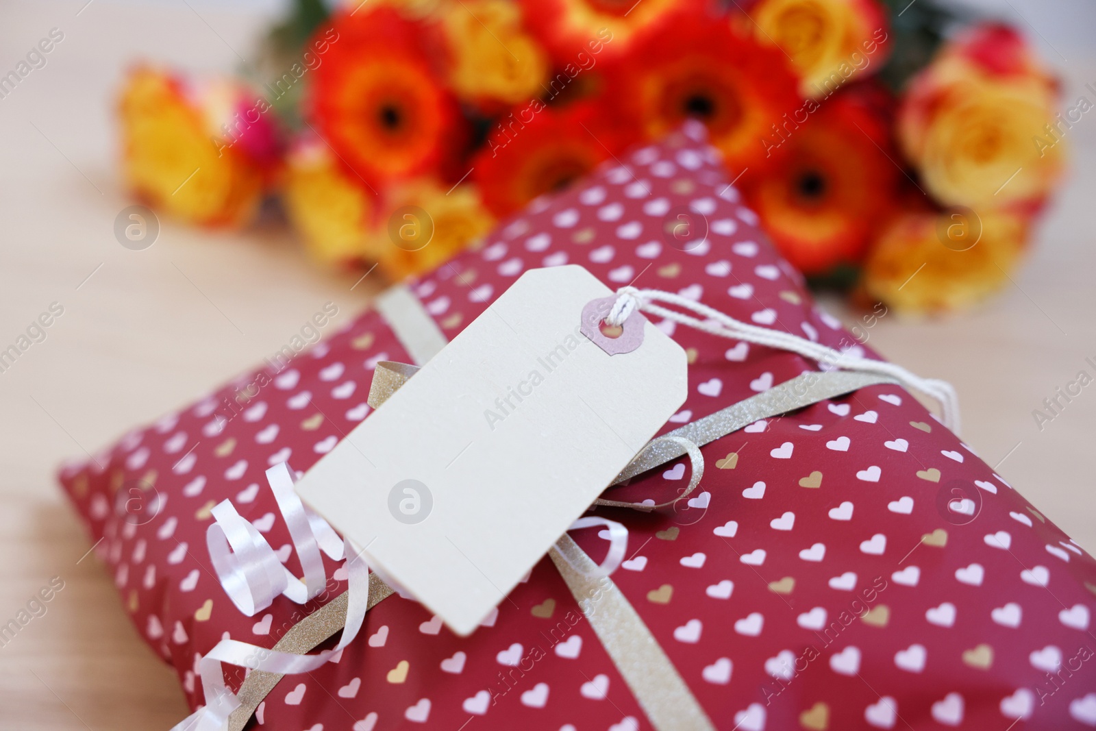 Photo of Parcel wrapped in heart patterned paper with cardboard tag and beautiful flowers on table, closeup. Space for text