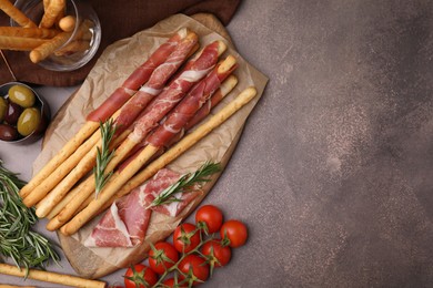 Photo of Delicious grissini sticks with prosciutto and snacks on brown table, flat lay. Space for text
