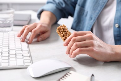 Photo of Woman holding tasty granola bar working with computer at light table in office, closeup